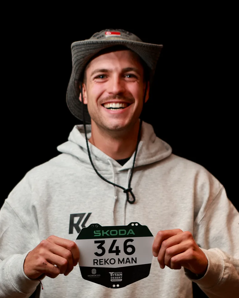 Xavier Miquel, con sudadera clara y sombrero de ala, sostiene el dorsal 346 de REKO MAN en la Titan Desert